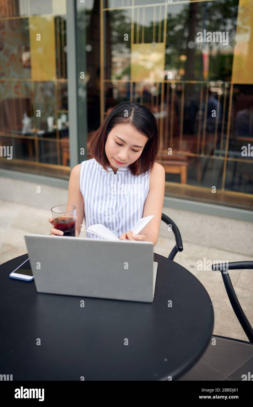 Belle confiance woman in casual clothing reading book et de boire du café tout en restant assis dans un café avec un ordinateur portable et ordinateur portable Banque D'Images