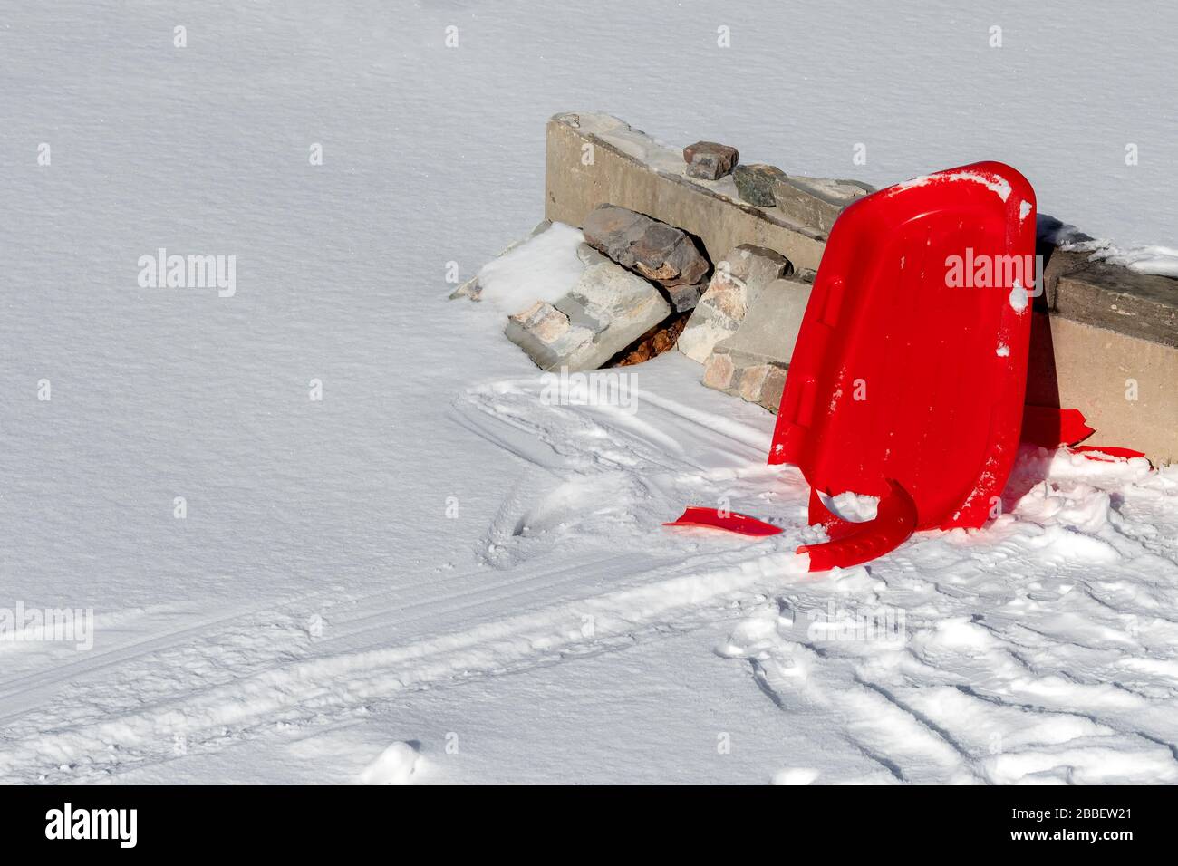 Un traîneau en plastique rouge cassé à l'extrémité du sentier qu'il a laissé dans la neige. Il penche contre un mur de béton, et des pièces sont sur le sol. Beaucoup de ro Banque D'Images