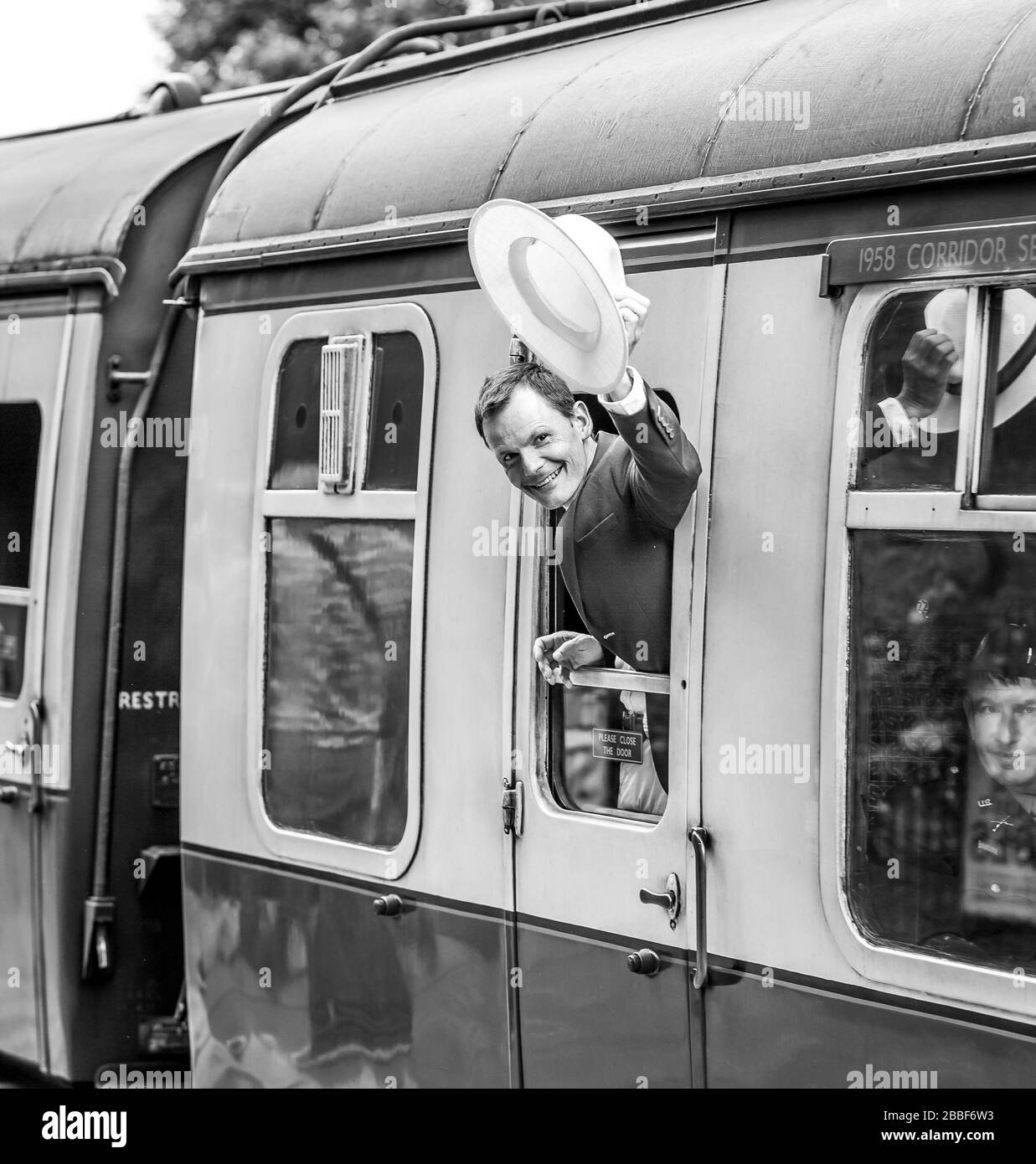 Noir et blanc, souriant homme des années 1940 à bord d'un train à vapeur britannique, penché en dehors de la fenêtre de calèche de chemin de fer d'époque agitant Au revoir avec chapeau, chemin de fer de la vallée de Severn. Banque D'Images