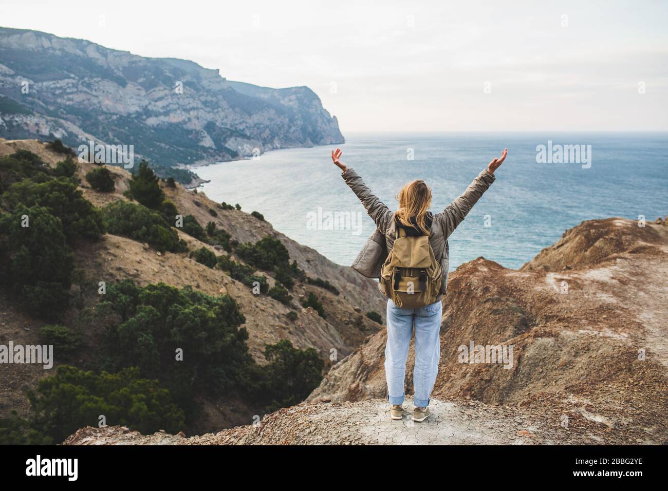 Backpacker hippie femme heureux de profiter d'une vue imprenable sur la mer et la montagne depuis la colline. Vue de derrière. Concept de voyage. Personne non reconnaissable dans un Jean et un sac à dos beige. Banque D'Images