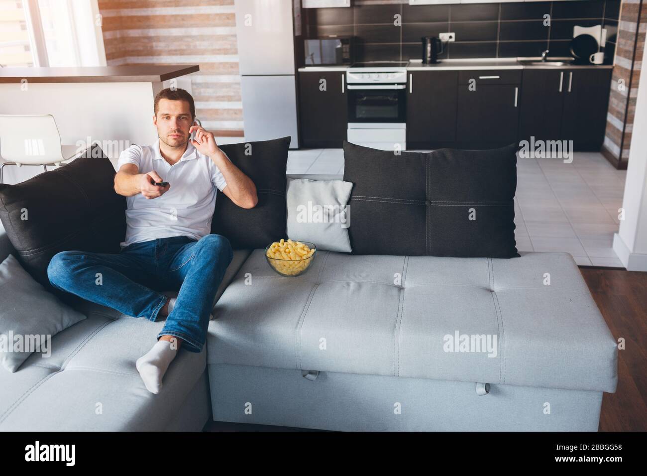 Jeune homme regarder la télévision dans son propre appartement. Un gars occupé assis sur le canapé et parler au téléphone. Changement de chaînes tv avec télécommande. Divertissez-vous Banque D'Images