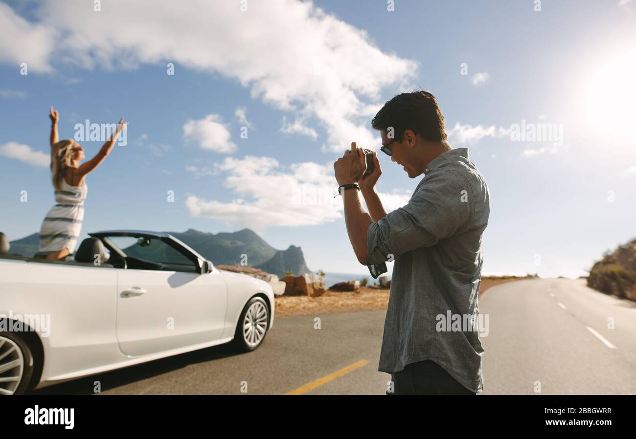 Homme avec appareil photo prendre des photos d'une femme debout dans un cabriolet. Femme debout avec ses mains soulevées à l'intérieur d'une voiture avec son petit ami prenant son pH Banque D'Images