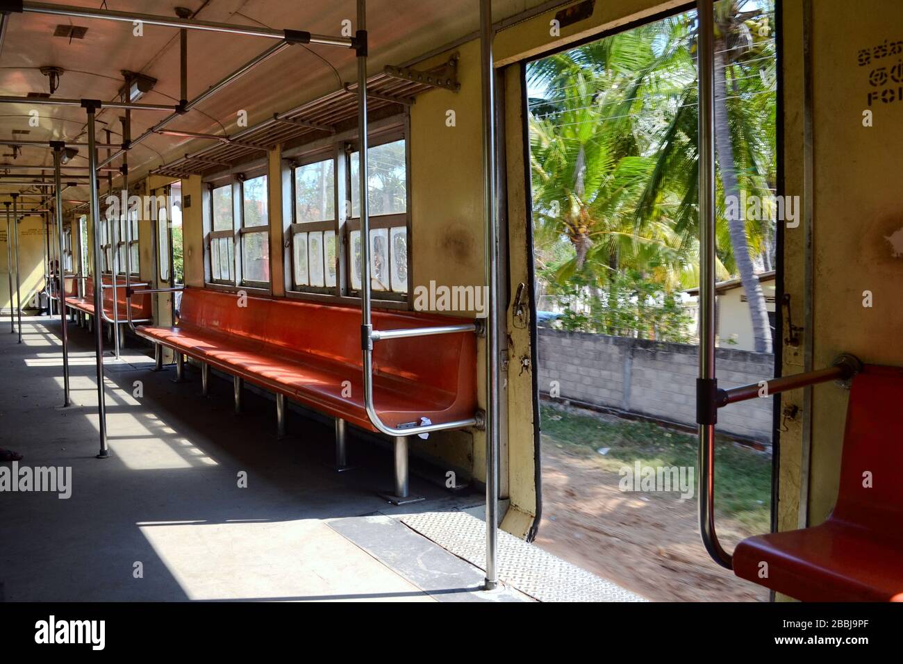 Intérieur d'un train de banlieue vide en déplacement. Palmiers tropicaux à travers portes ouvertes. Sièges durs orange sans personne ni mains courantes en métal. Sri Lanka Banque D'Images