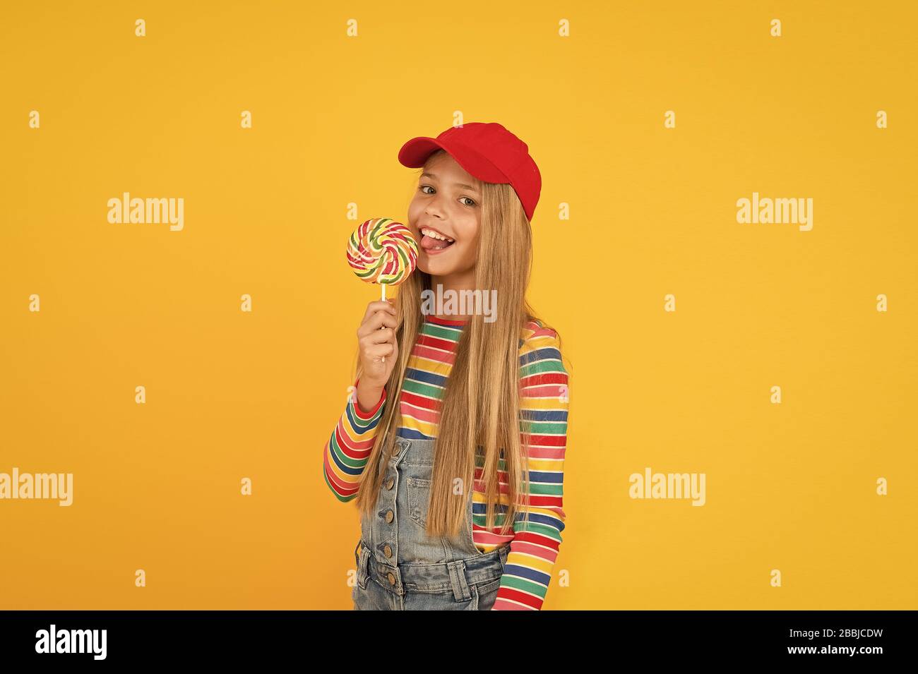 Lucette la rend heureuse. Cute little girl eating candy swirl arc-en-ciel sur fond jaune. Petit enfant léchant des bonbons durs sur stick. Adorable happy kid profitant de sucre candi dessert. Magasin de bonbons. Banque D'Images