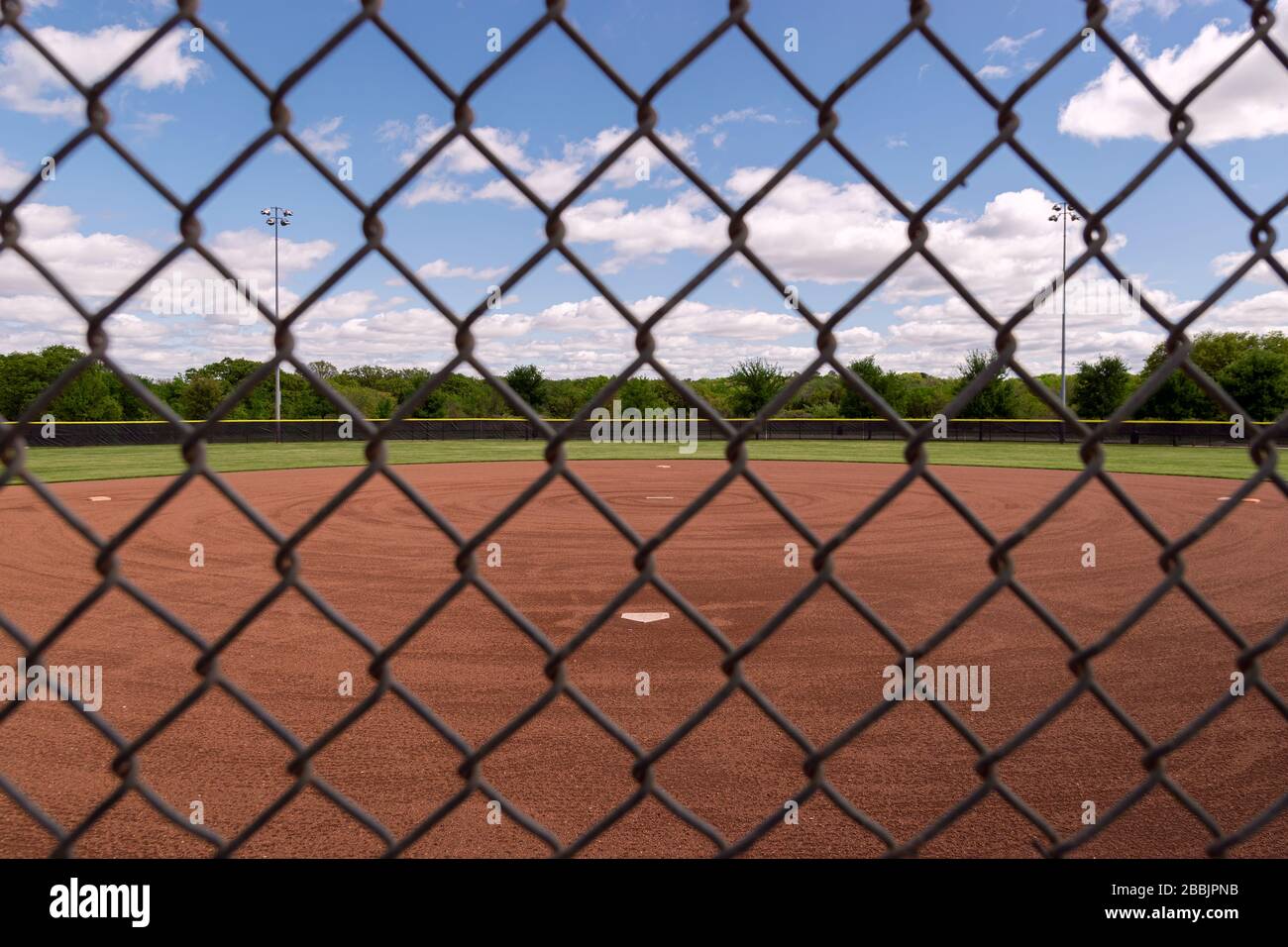 Regardez un terrain de base-ball à travers la grille à motifs en losange d'une clôture de maillon de chaîne de derrière la plaque d'accueil lors d'une belle journée de printemps avec partiellement le clo Banque D'Images