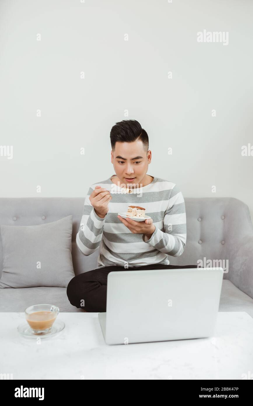 Jeune homme asiatique mangeant des gâteaux lorsqu'il est assis sur un canapé et à l'aide d'un ordinateur portable. Travailler à domicile. Banque D'Images