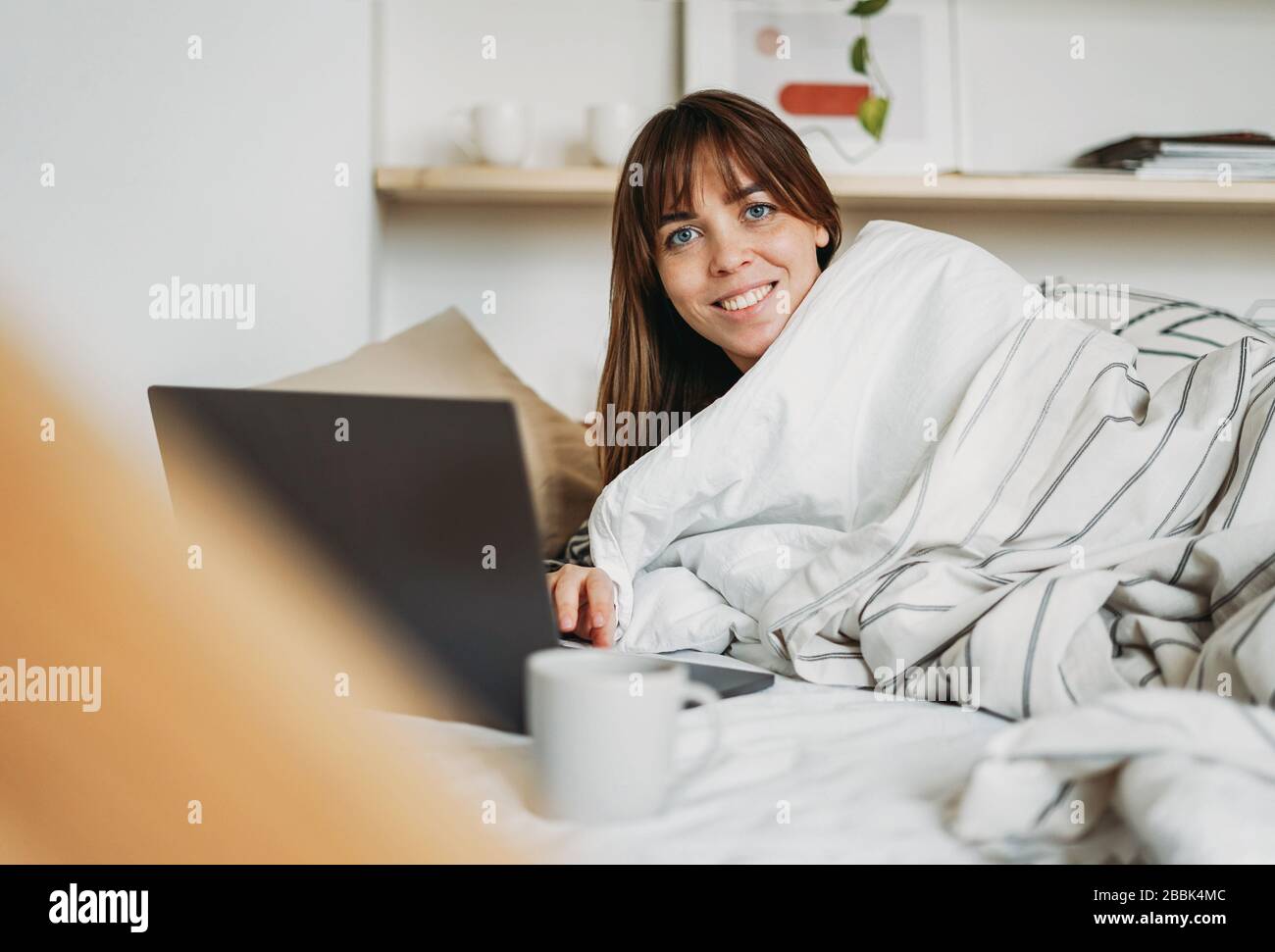 Travail jeune femme brunette freelance dans des vêtements décontractés avec ordinateur portable et tasse de thé réveil dans le lit à la maison dans un séjour intérieur lumineux à la maison. Banque D'Images