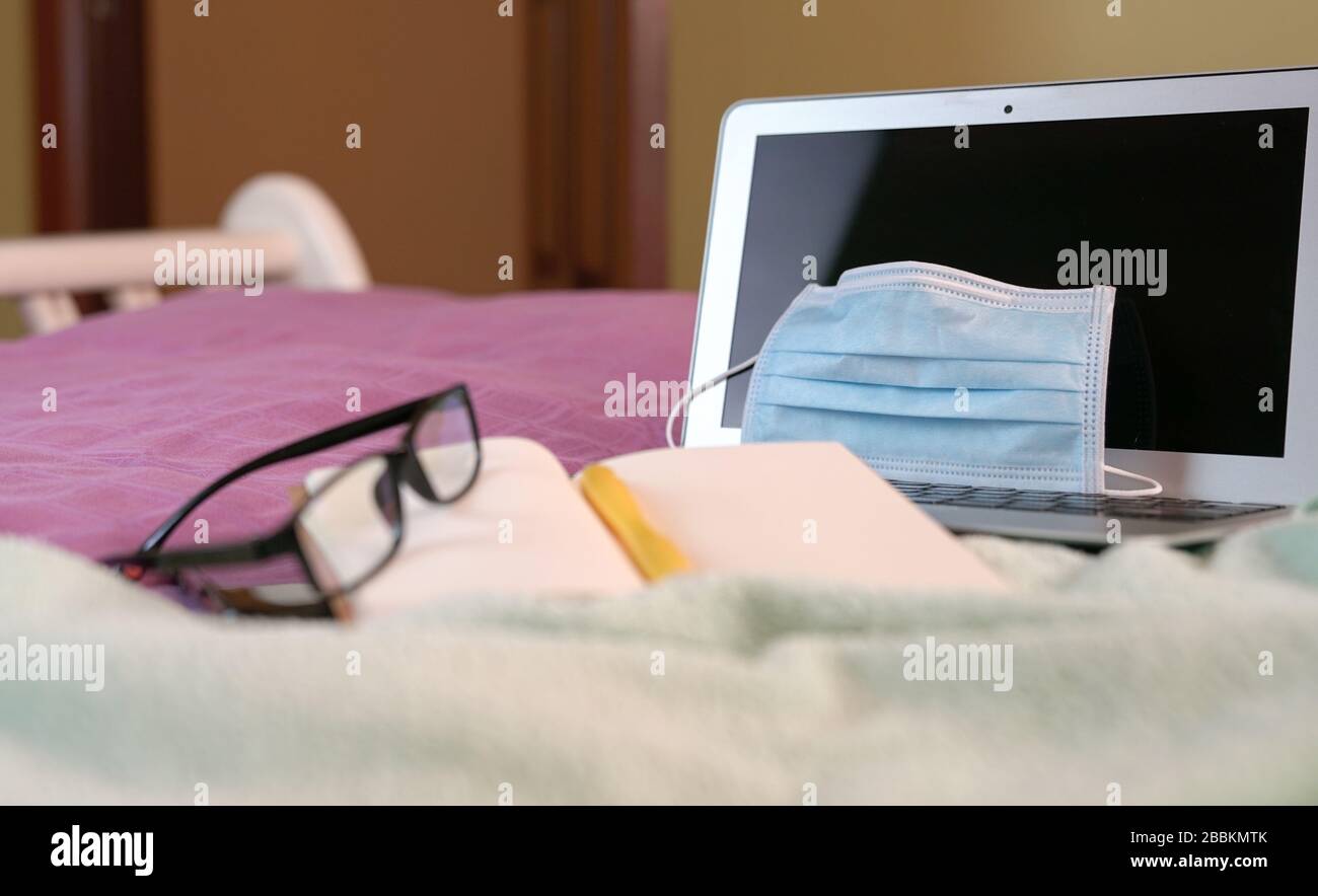 Espace de travail à la maison avec ordinateur portable, bloc-notes et lunettes sur le lit pendant l'éclosion de coronavirus. Concentrez-vous sur le masque chirurgical. Banque D'Images