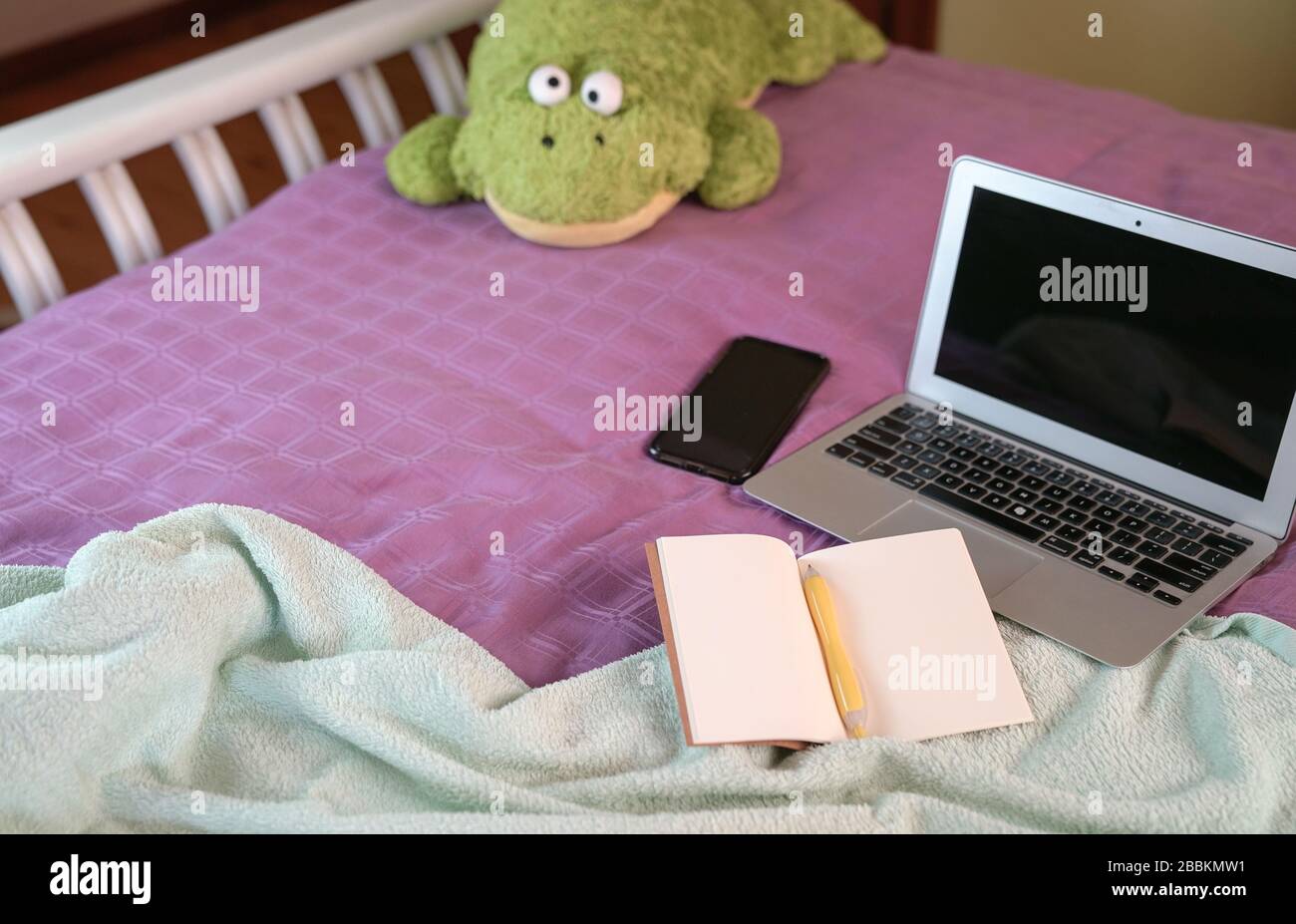 Ordinateur portable avec bloc-notes, stylo, téléphone portable et jouet doux sur le dessus du lit dans la chambre. Travailler à la maison. Banque D'Images