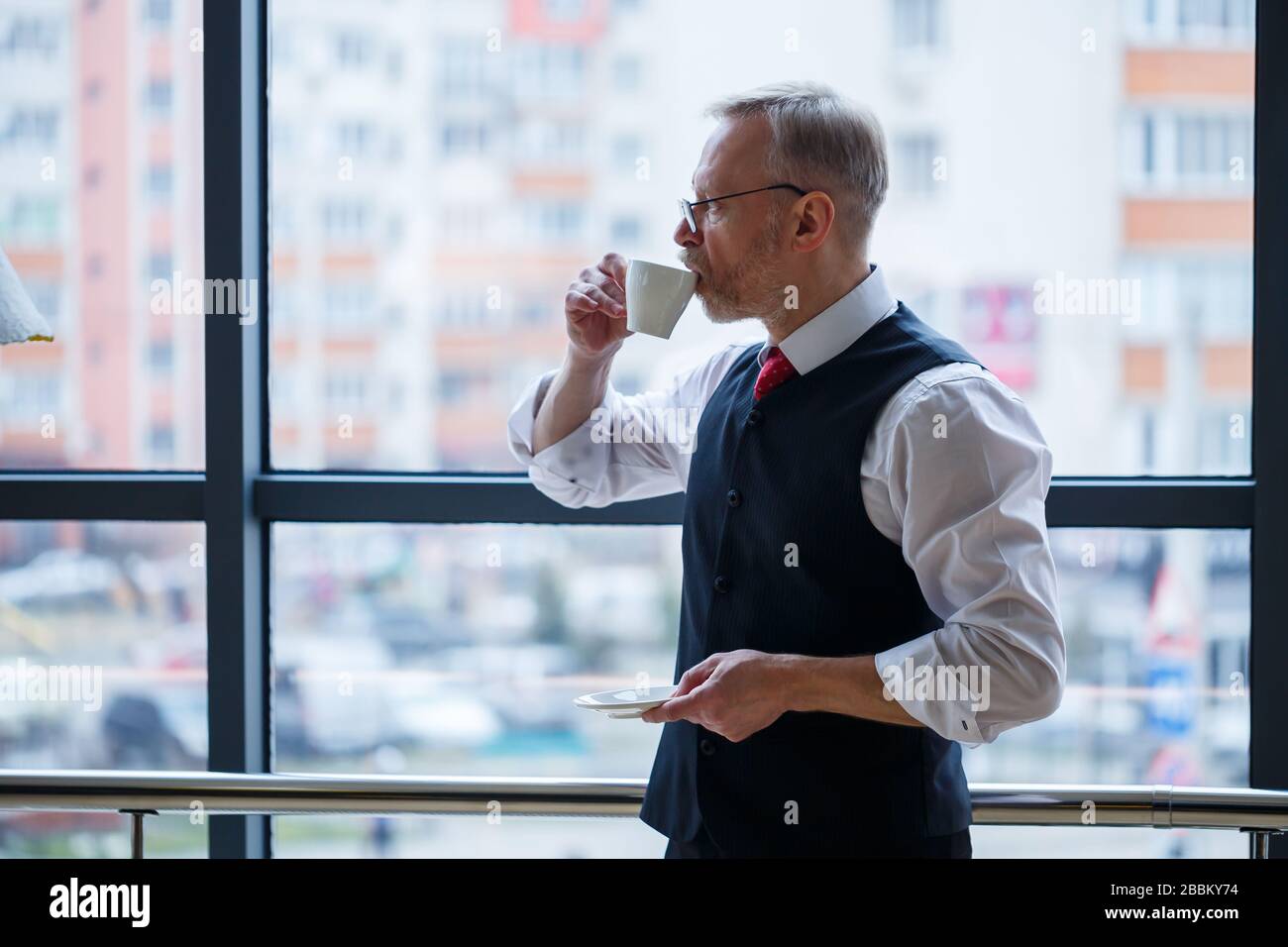 Sourire heureux directeur général pense à son développement de carrière réussi tout en se tenant avec une tasse de café dans sa main dans son bureau près de la Banque D'Images
