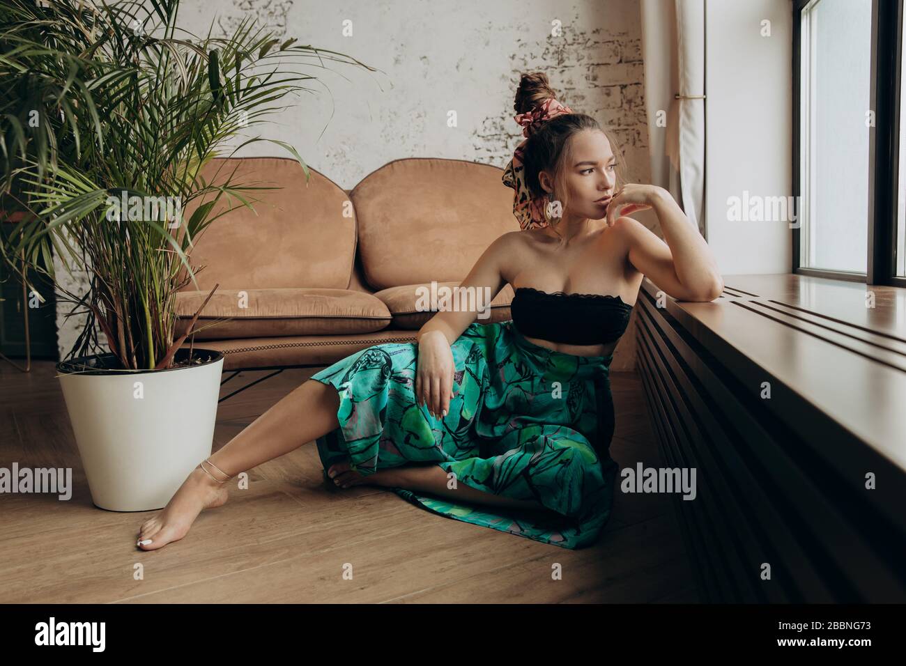 Une fille dans une robe tropicale orientale est assise près de la fenêtre. Les cheveux des gri sont rassemblés dans un grand pain. Banque D'Images