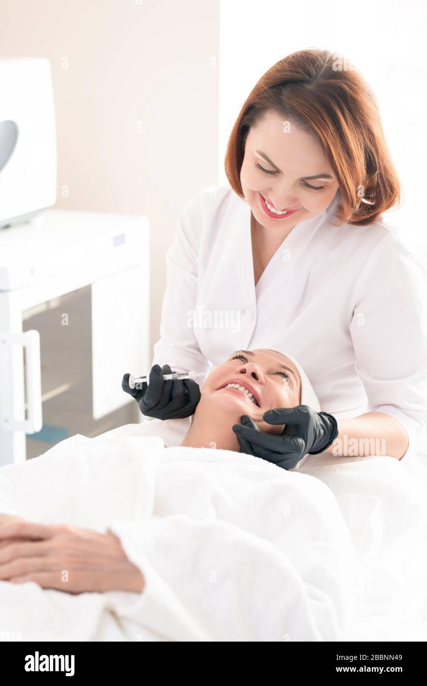 Professionnel esthétique souriant en gants chirurgicaux et blouse de laboratoire injectant la soutireuse dans le visage pour éliminer les rides de la femme mûre Banque D'Images