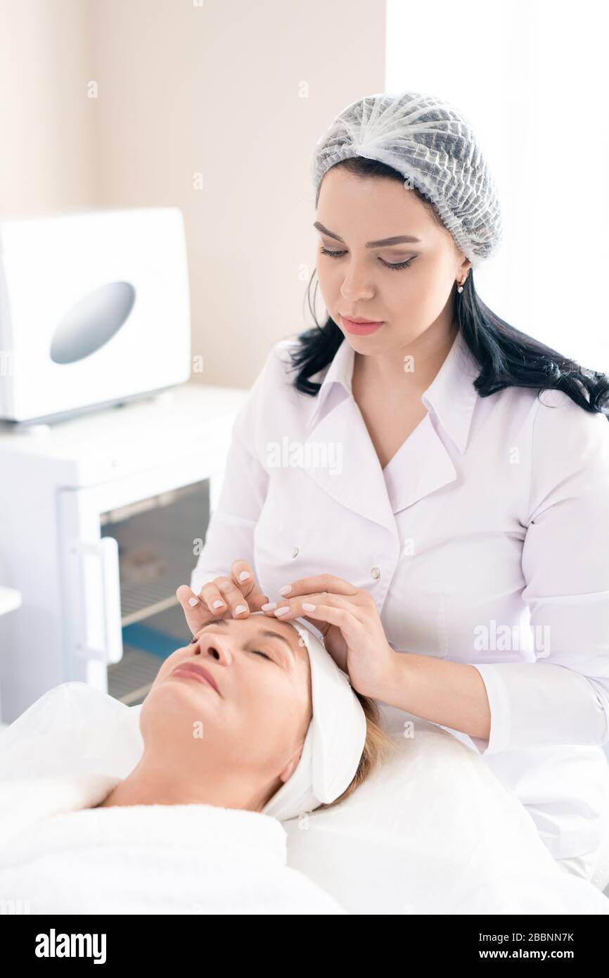 Professionnel de la beauté en bouchon chirurgical faisant un massage relaxant du visage à la maturité client pour lisser les rides Banque D'Images