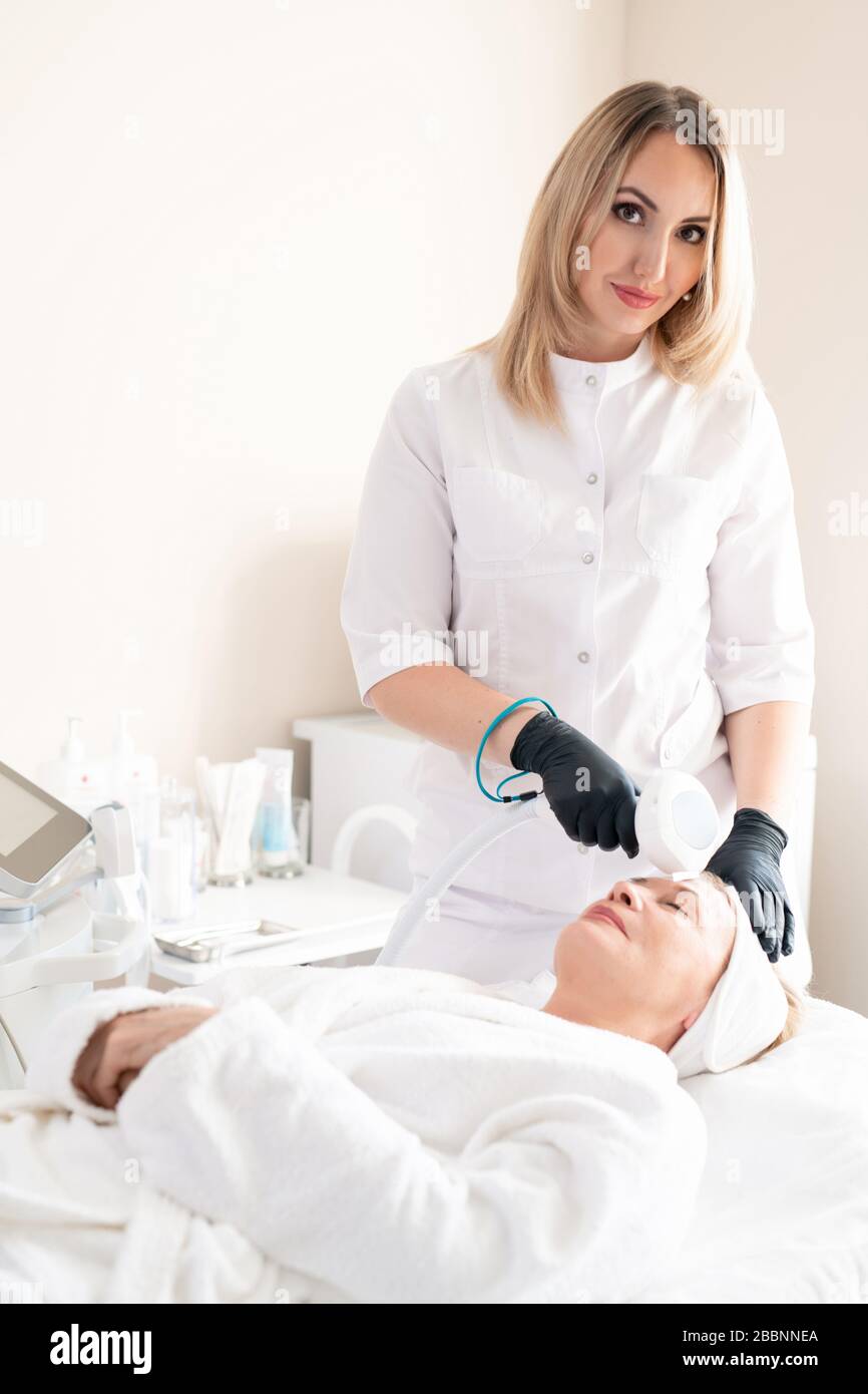 Portrait du contenu jeune cosmologiste dans des gants chirurgicaux utilisant un appareil de beauté pour nettoyer la peau de la femme mûre Banque D'Images