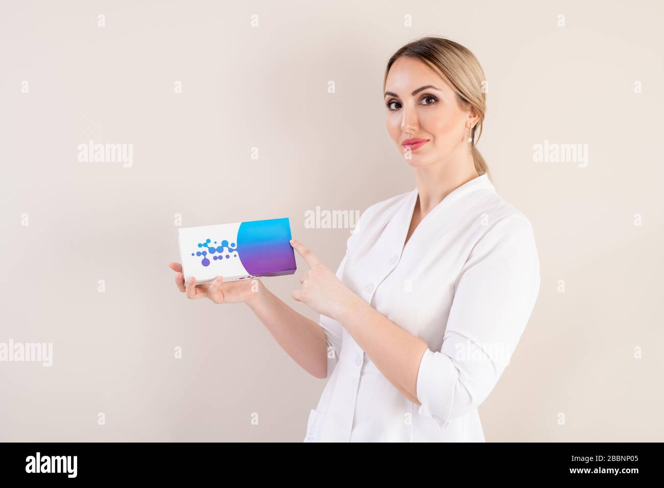 Portrait du contenu jeune cosmologiste féminin en blouse de laboratoire recommandant un produit de remplissage sur fond isolé Banque D'Images