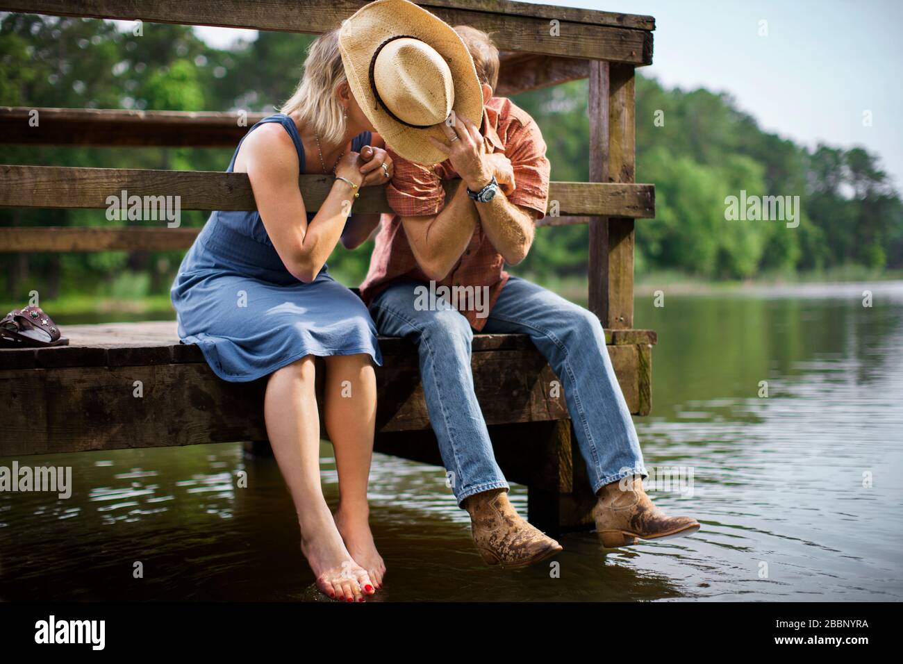 L'homme mûr tient un chapeau de cowboy pour bloquer la vue pendant qu'il  penche pour donner à sa femme un baiser romantique lorsqu'ils s'assoient  ensemble sur une jetée en bois au-dessus d'un
