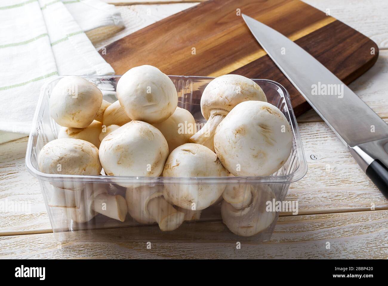 Tas de champagnes crus entiers dans un plateau alimentaire en plastique, planche à découper et couteau sur une table en bois. Champignons en tant que protéines végétales, alimentation alimentaire brute. Banque D'Images