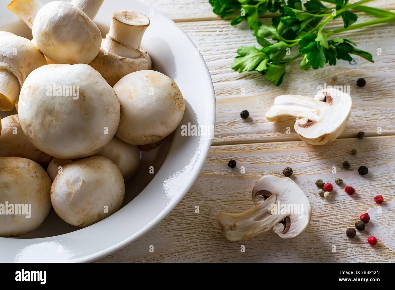 Tas de champagnes frais crus entiers dans un bol blanc, bouquet de persil et de poivre sur une table en bois. Champignons en tant que protéines végétales, alimentation alimentaire brute Banque D'Images