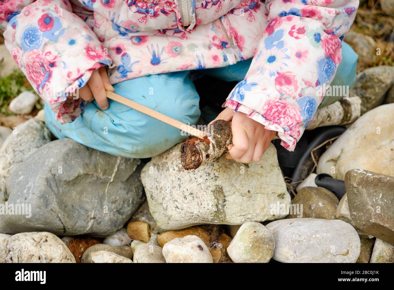 Gros plan de la partie basse de la fille caucasienne de 4 ans dans des vêtements chauds assis sur des pierres dans le jardin et jouant avec deux bâtons en bois. Je l’ai vu Banque D'Images