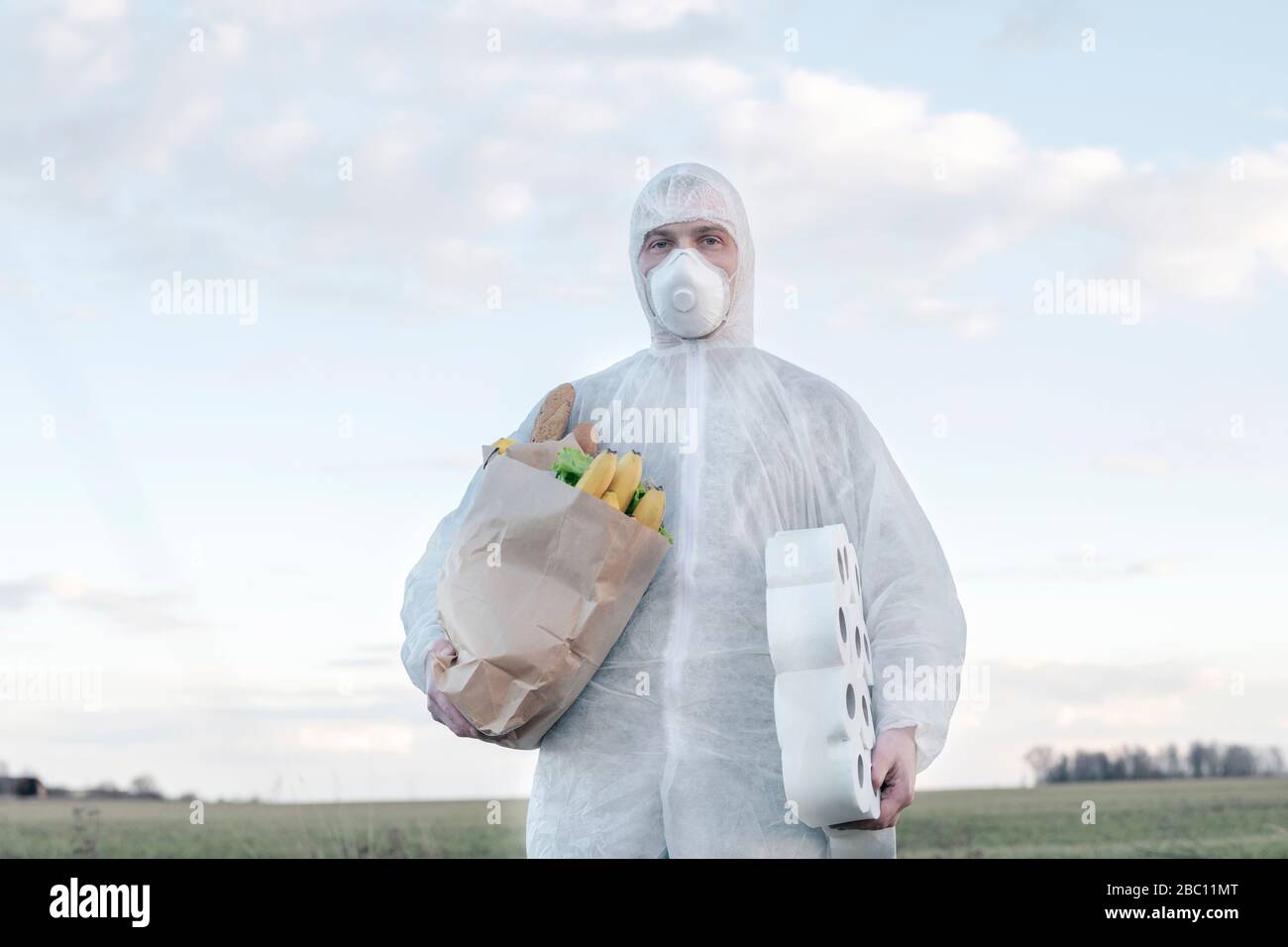 Homme portant un costume de protection et un masque contenant des rouleaux de toilette et un sac d'épicerie à la campagne Banque D'Images