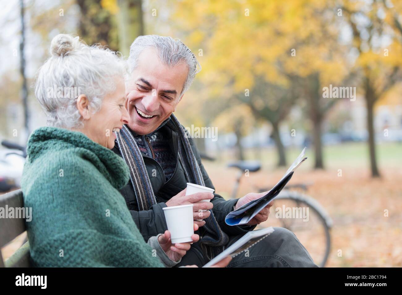 Couple senior souriant lisant le journal et buvant du café sur banc dans le parc d'automne Banque D'Images