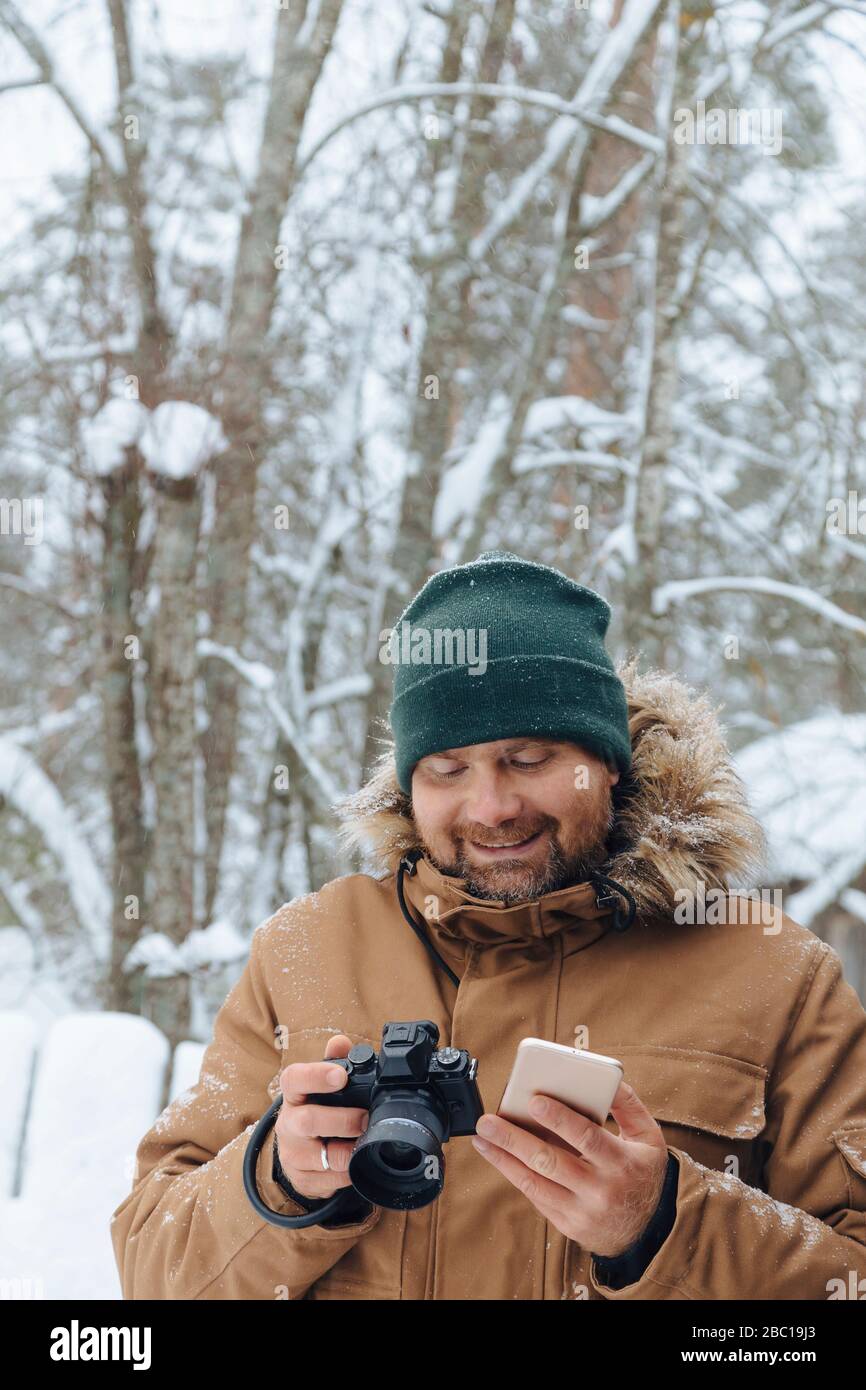 Portrait de l'homme détendu avec appareil photo numérique et téléphone portable en forêt d'hiver Banque D'Images