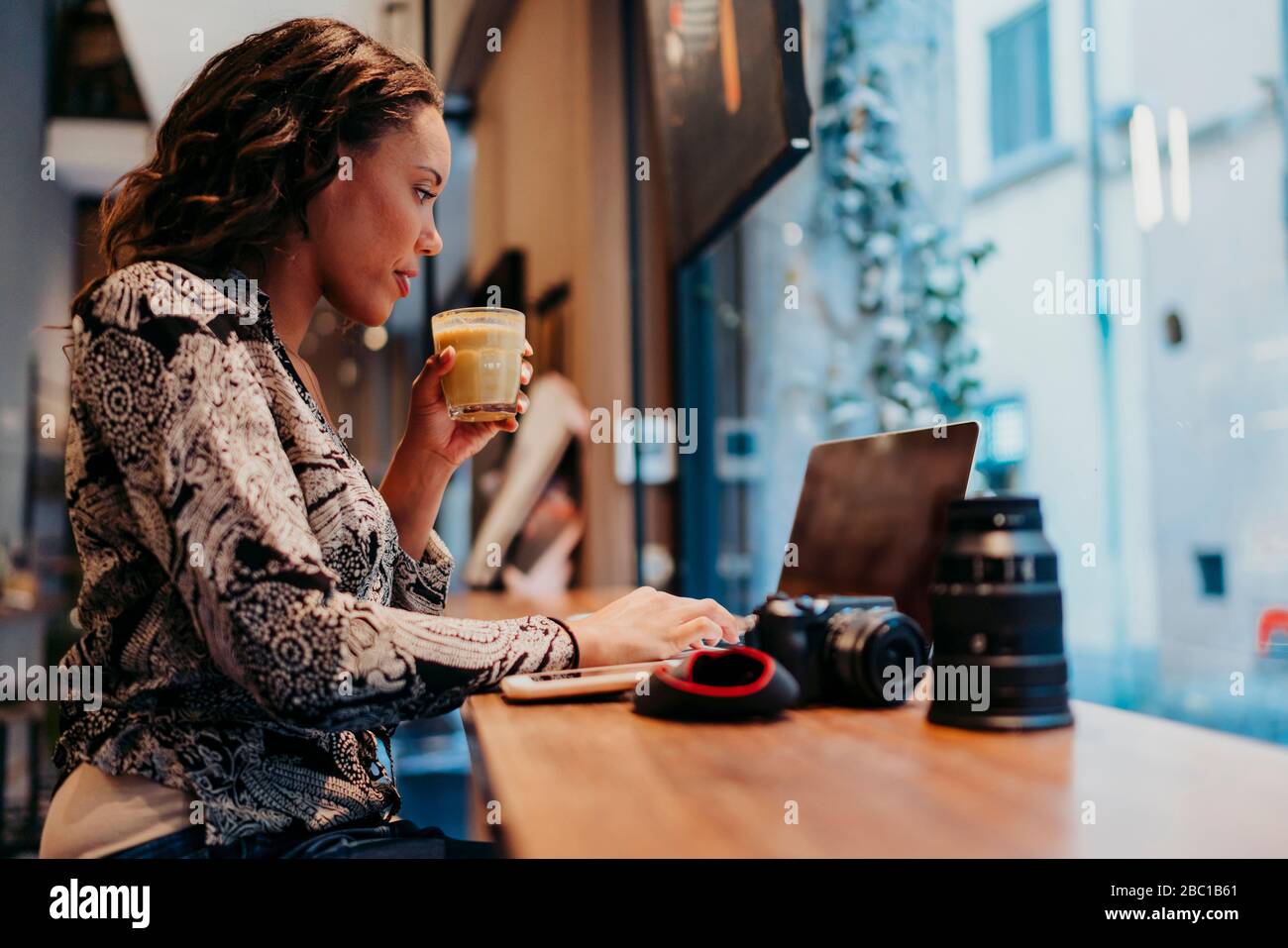 Jeune femme avec un appareil photo utilisant un ordinateur portable dans un café derrière le panneau de fenêtre Banque D'Images