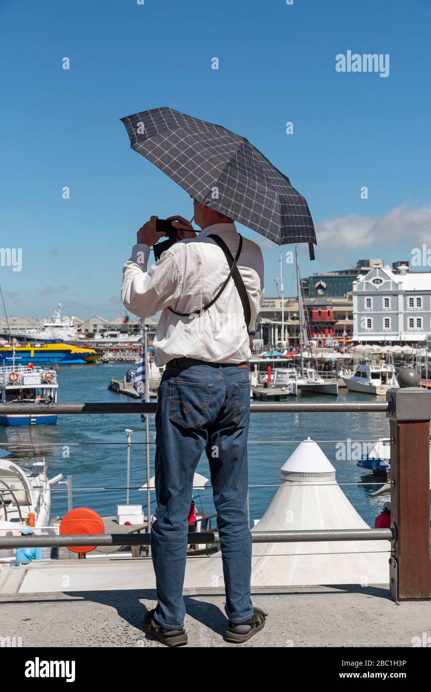 Le Cap, Afrique du Sud. 2019. Homme tenant un parasol et utilisant un appareil photo pour prendre une photo du bord de mer. Banque D'Images