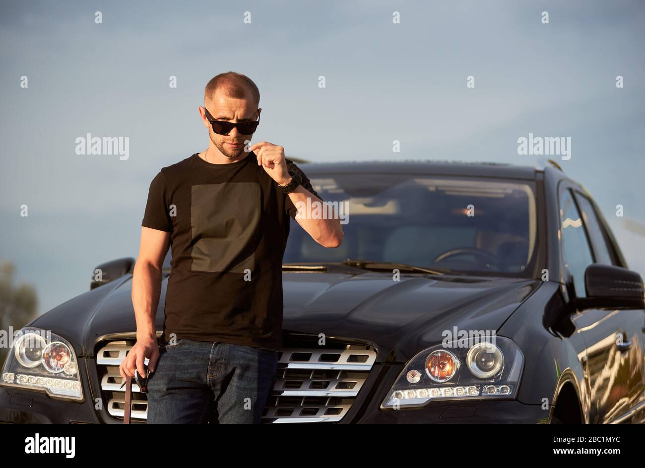 Portrait d'un homme plein de confiance en soi, debout devant sa jolie voiture noire, portant des lunettes de soleil noires, un t-shirt noir et un Jean, gros plan Banque D'Images