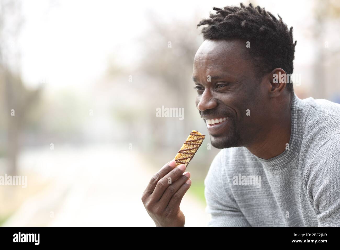Un homme noir heureux mangeant un bar à céréales dans un parc donnant sur l'extérieur Banque D'Images