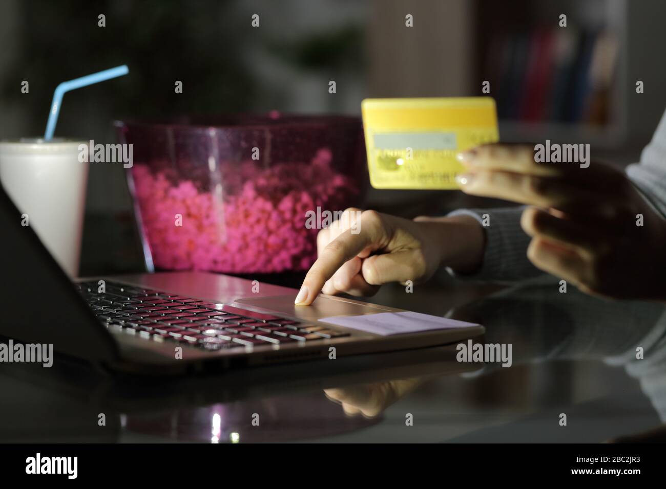 Gros plan de fille payant main en ligne avec carte de crédit sur ordinateur portable la nuit avec pop-corn bol et soda à la maison Banque D'Images