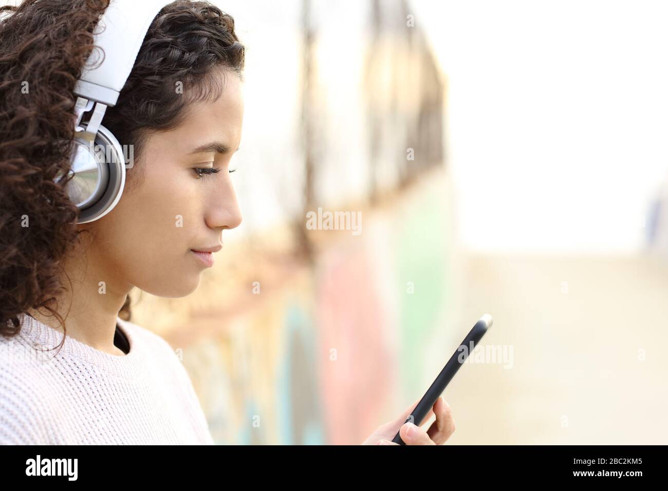 Vue latérale portrait d'une jeune fille latina sérieuse écoutant de la musique portant des écouteurs et regardant le contenu de smartphone dans la rue Banque D'Images