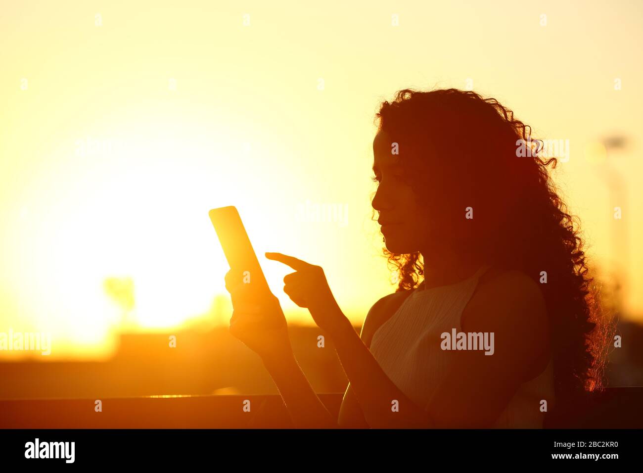 Silhouette d'une femme qui vérifie le téléphone mobile assis sur un banc au coucher du soleil Banque D'Images