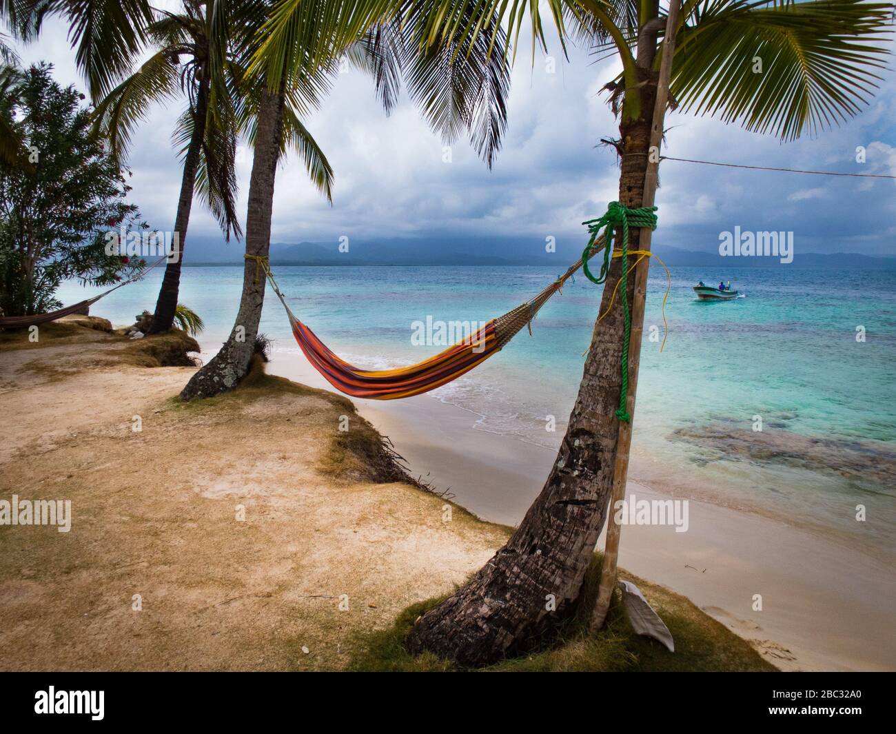 Un hamac se trouve entre deux palmiers à noix de coco au bord de l'eau sur une île de l'océan des Caraïbes. Un bateau de pêche approche à la distance. Banque D'Images