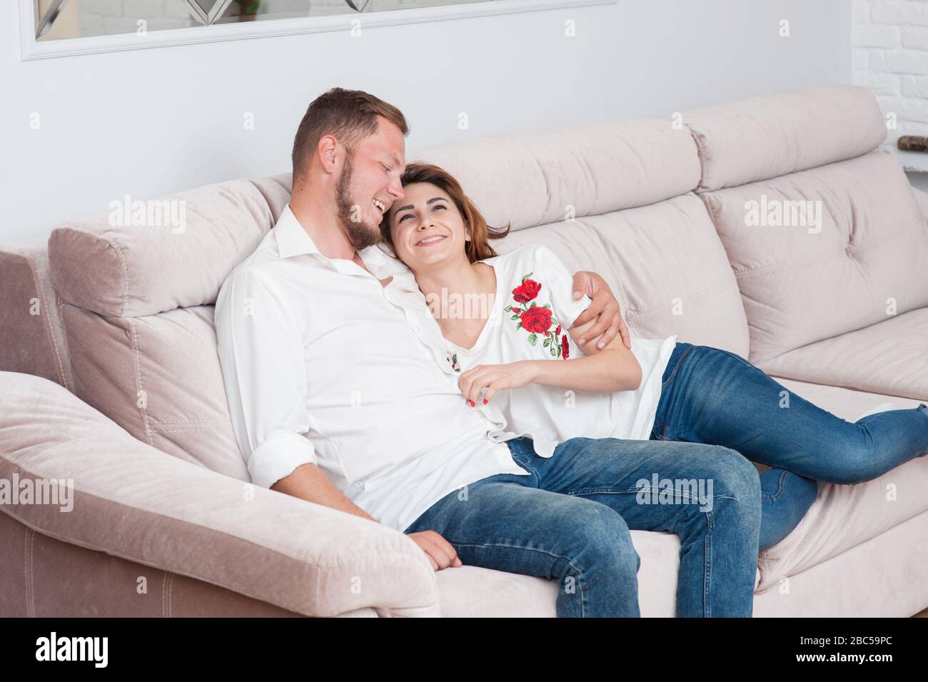 heureuse famille jeune de barbu homme et femme en chemise blanche et jeans bleu assis ensemble riant à l'intérieur à la maison pendant l'auto-isolation Banque D'Images