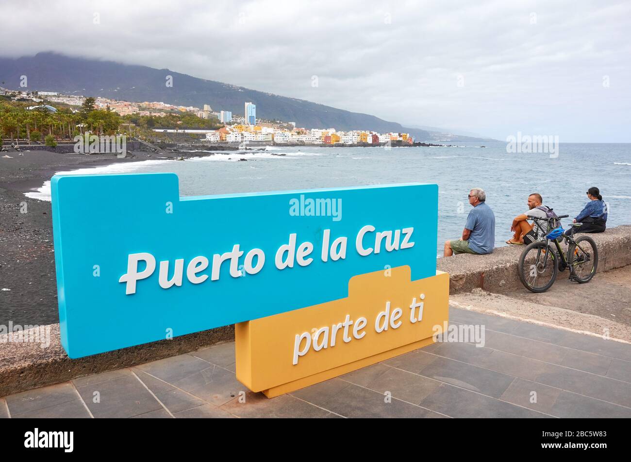Puerto de la Cruz, Tenerife, Espagne - 01 mai 2019: Les gens sont assis par un panneau accueillant donnant sur une plage vide en journée nuageux. Banque D'Images