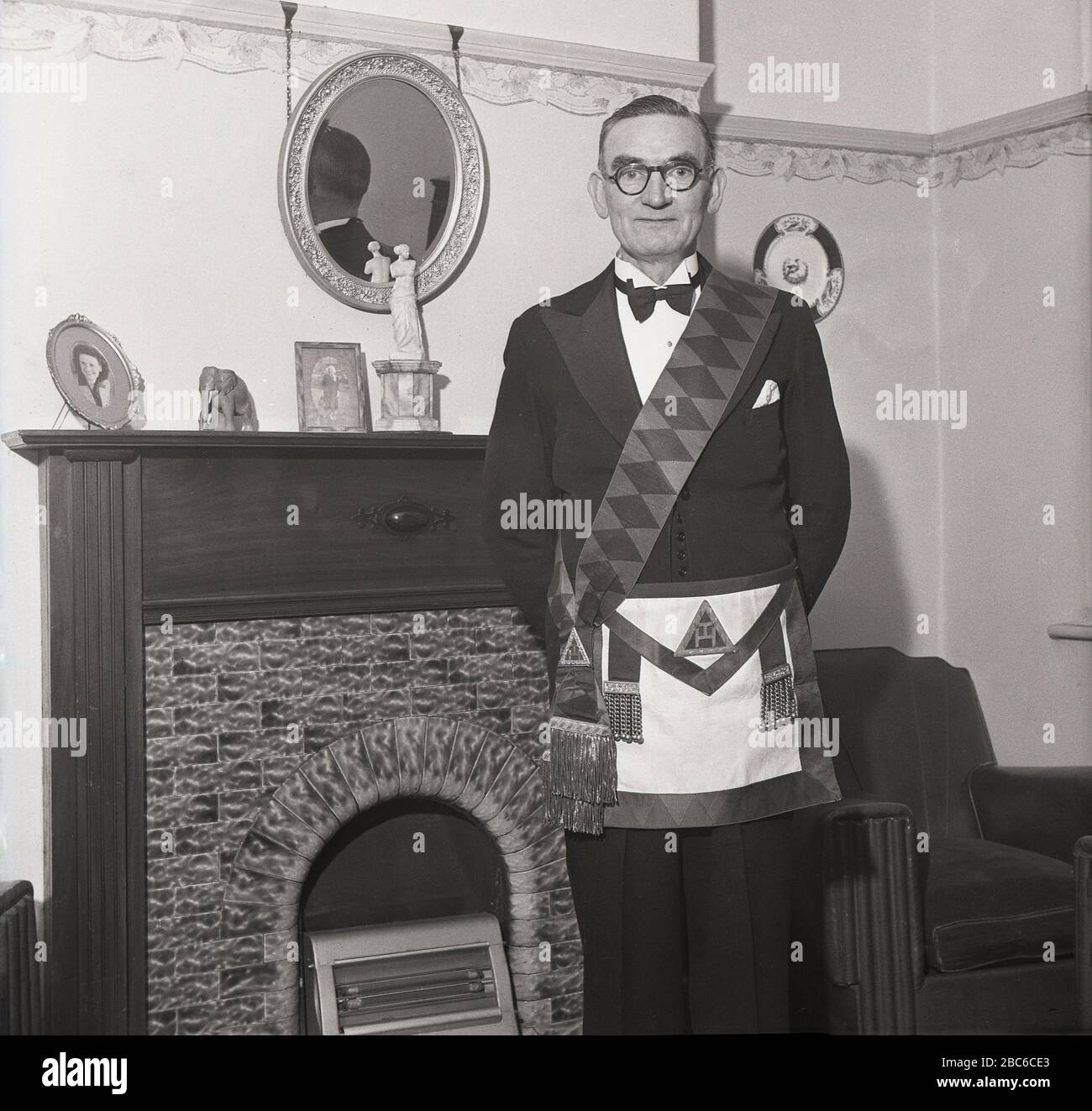 Années 1950, historique, debout fièrement près de la cheminée dans sa chambre avant, un freemason portant son tablier de robe ou regalia. Également connu sous le nom de tablier Craft Master Masons, il dérive du tablier de travail des anciens maçons en pierre et est considéré comme un emblème de l'innocence et de la pureté de la vie. Le tablier d'un maître maçon est normalement fabriqué à partir de peau d'agneau et, à l'époque primitive, il s'agissait d'une décoration ecclésiastique (religieuse). Banque D'Images