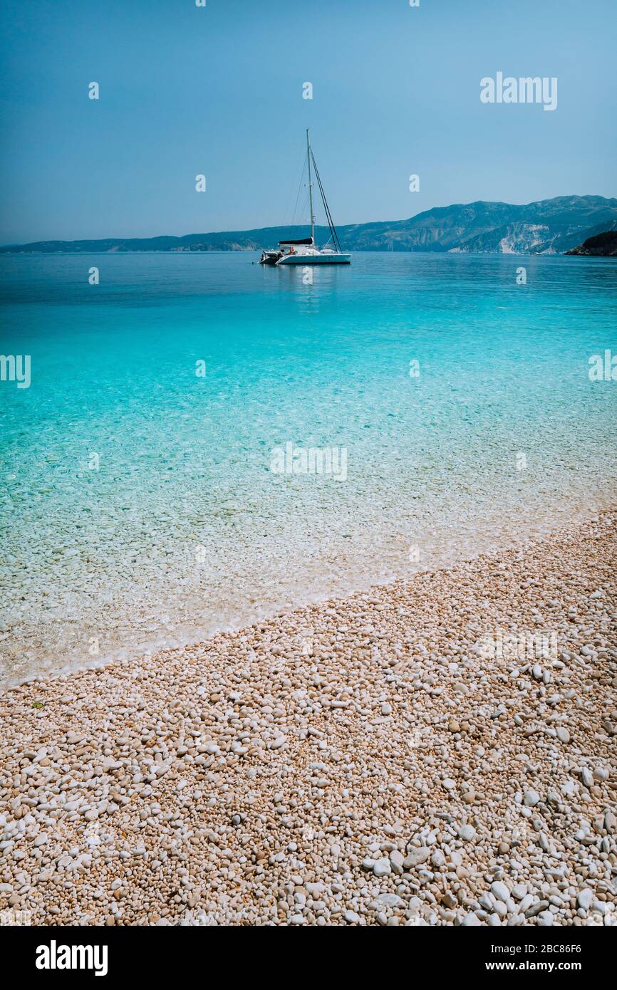Plage de galets avec eau bleue pure et claire azure et bateau à voile catamaran à l'ancre à l'horizon. Banque D'Images