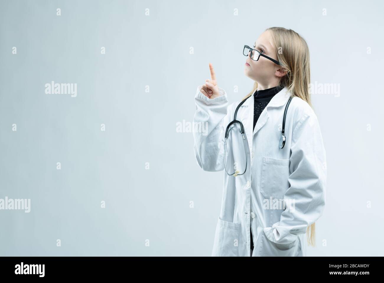 Petite fille vêtue comme infirmière ou médecin avec des lunettes de grande taille, un manteau blanc et un stéthoscope tenant son doigt vers l'espace vierge de copie sur blanc Banque D'Images