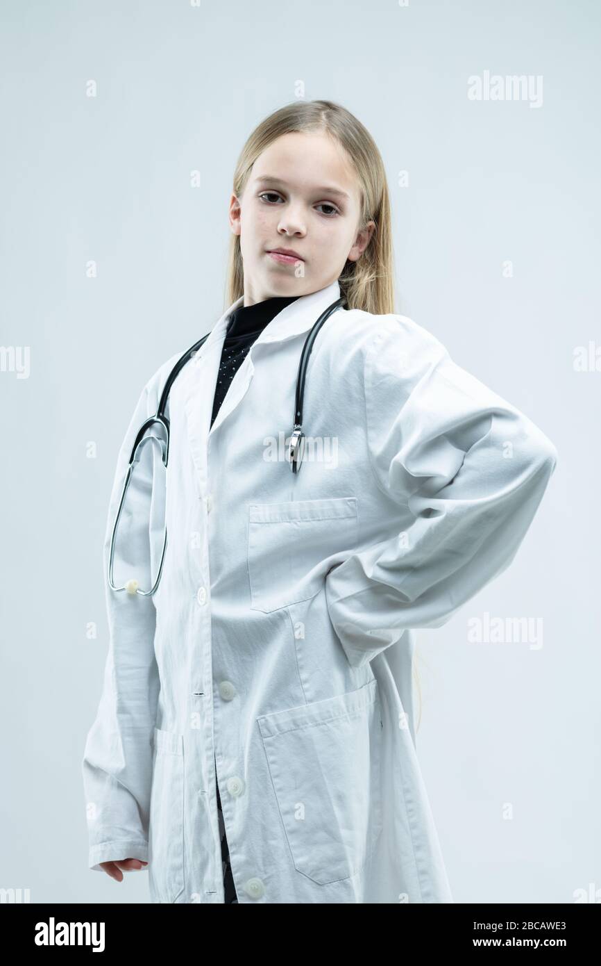 Fière jeune fille dans un manteau de laboratoire blanc jouant à être médecin ou infirmière avec stéthoscope autour de son cou isolé sur blanc Banque D'Images