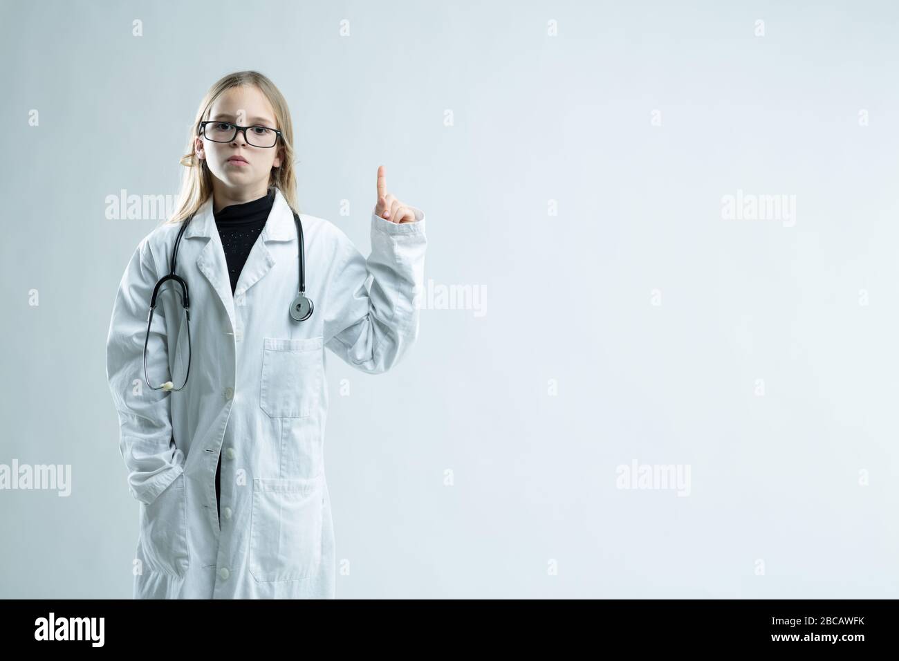 Jolie petite fille jouant à être un médecin debout dans un manteau blanc de laboratoire avec stéthoscope et de grands verres tenant son doigt isolé sur la neige Banque D'Images