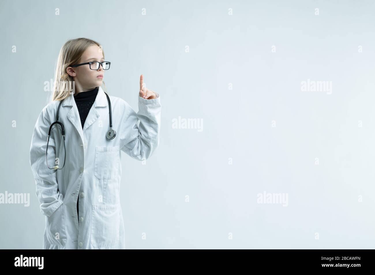 Jolie petite fille blonde jouant à être médecin dans un manteau de laboratoire et stéthoscope tenant son doigt regardant vers l'espace vierge de copie sur blanc Banque D'Images