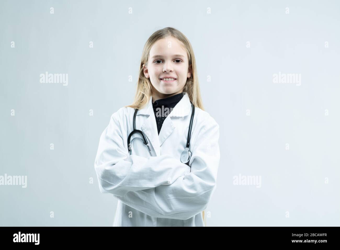 Sourire sympathique petite fille vêtue comme médecin dans un manteau blanc de laboratoire avec stéthoscope debout avec bras repliés à l'appareil photo sur blanc avec c Banque D'Images