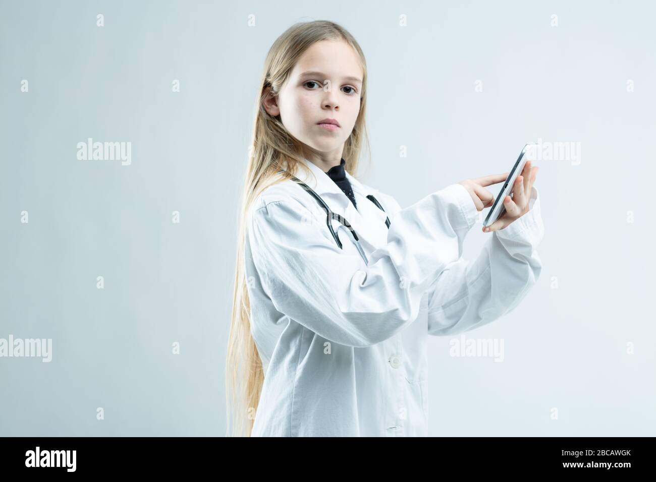 Jeune fille sérieuse vêtue comme médecin ou infirmière tenant un téléphone mobile dans ses mains lorsqu'elle se tourne vers la stare à l'appareil photo isolé sur blanc Banque D'Images