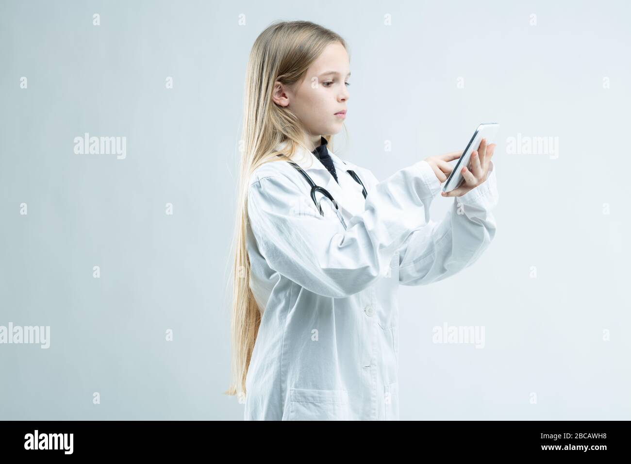 Jeune fille vêtue d'un manteau de laboratoire médical avec stéthoscope prétendant être médecin debout en utilisant son téléphone portable isolé sur blanc Banque D'Images