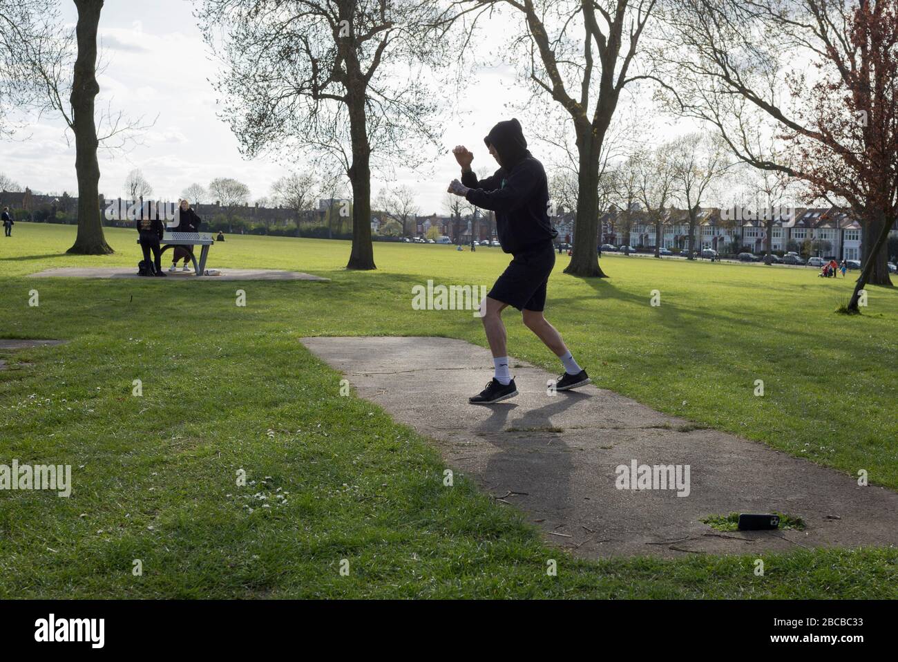 Alors que la deuxième semaine du maintien du Coronavirus se poursuit et une semaine avant Pâques, lorsque le Premier ministre Boris Johnson rappelle aux Britanniques de rester localement et de ne pas se rendre dans des lieux de beauté, le nombre de morts au Royaume-Uni atteint 2 921, avec 1 million de cas de Covid-19 dans 181 pays. Michial, un poids moyen de Brooklyn spars avec une application de boxe sur son téléphone en après-midi soleil et les températures printanières à Ruskin Park à Herne Hill, 3 avril 2020, dans le sud de Londres, Angleterre. Banque D'Images