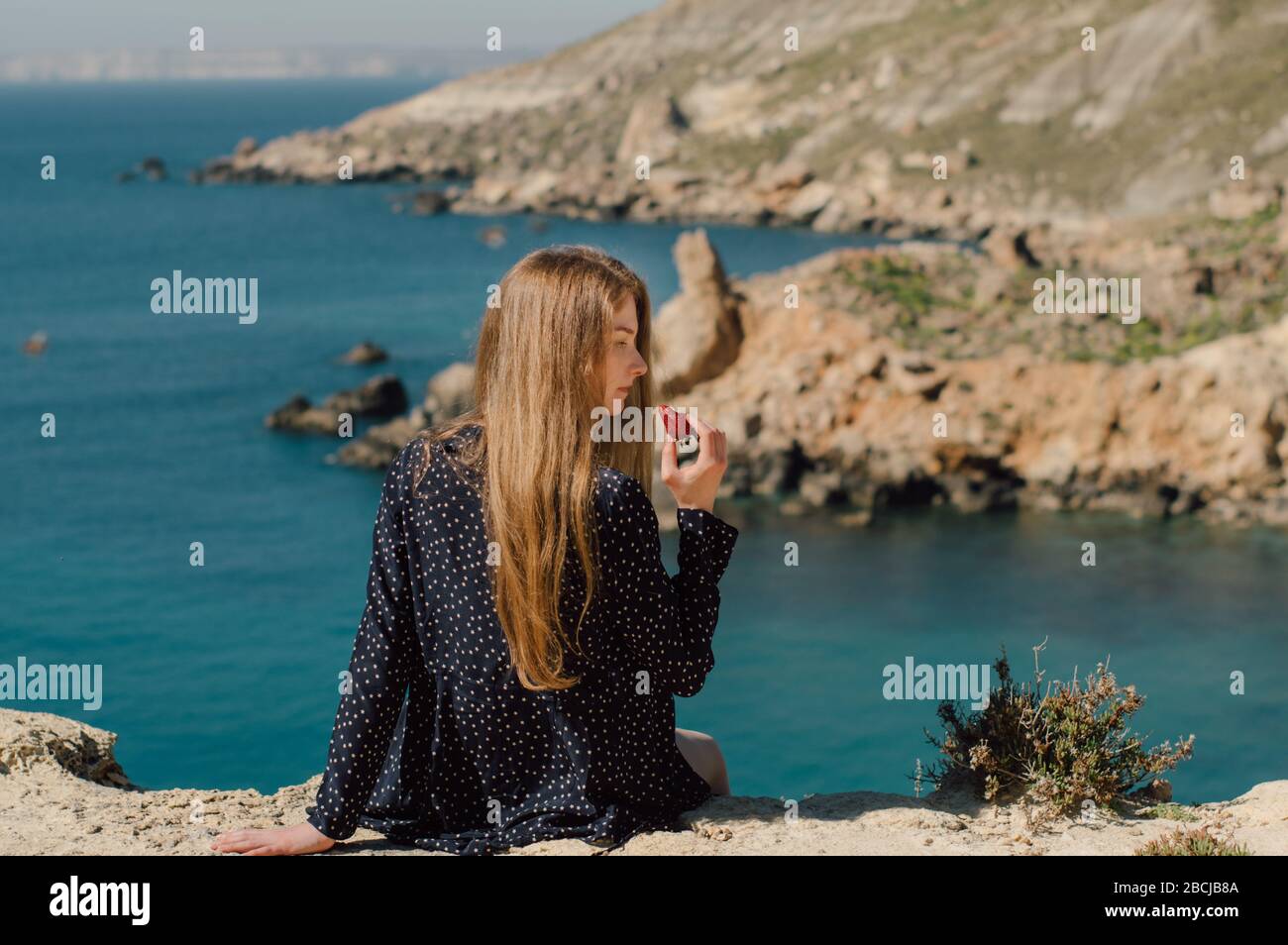 Belle fille blonde avec fraise en main assise sur le bord de la montagne avec vue sur la mer, vue latérale Banque D'Images