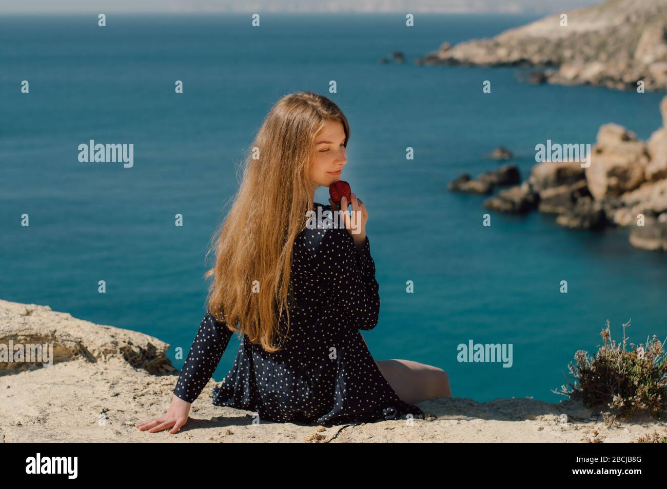 Belle fille blonde souriante avec fraise à la main assise sur le bord de la montagne avec vue sur la mer, vue latérale Banque D'Images