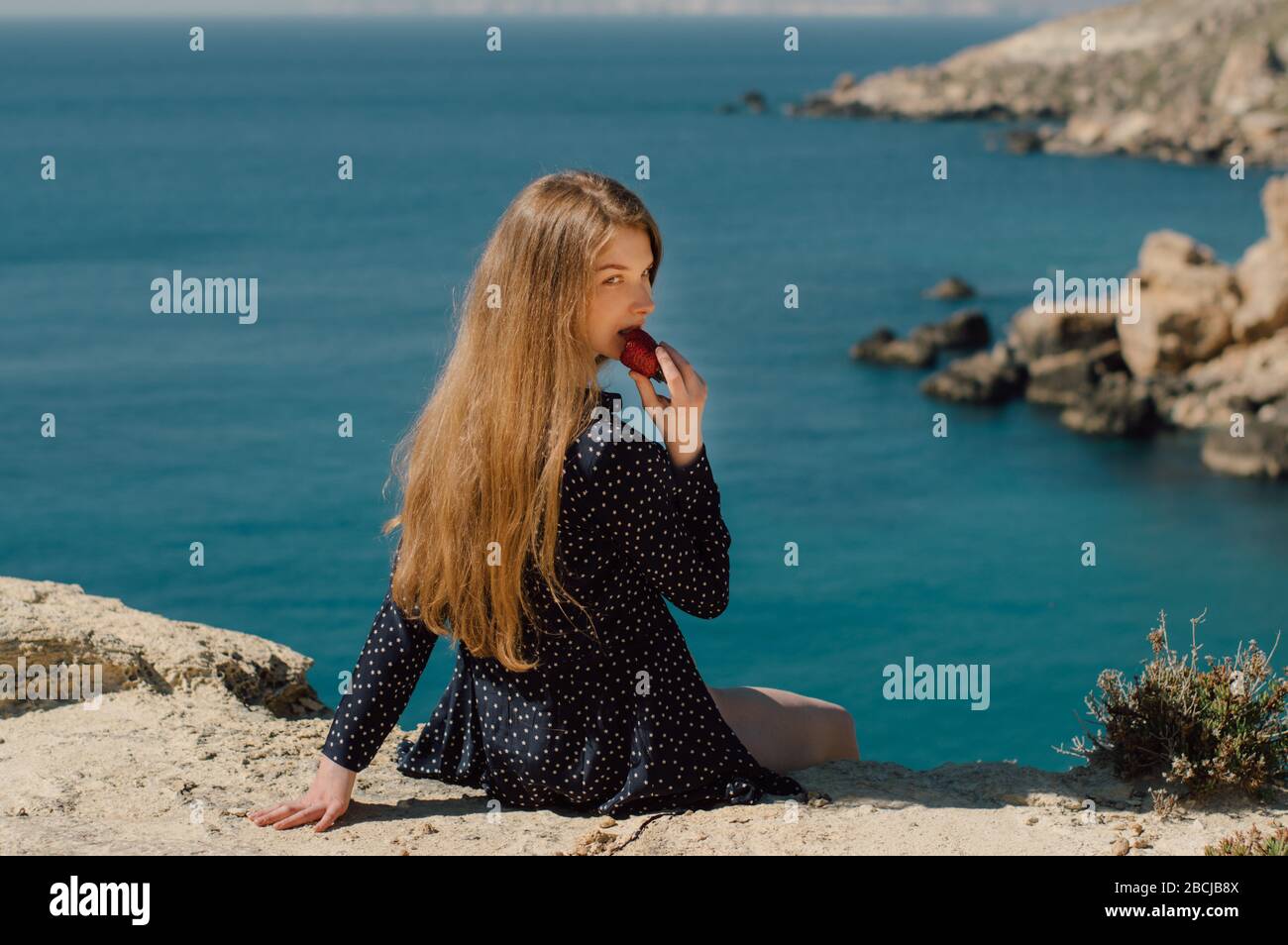 Belle fille blonde souriante mangeant fraise et assis sur le bord de la montagne avec vue sur la mer, vue latérale Banque D'Images
