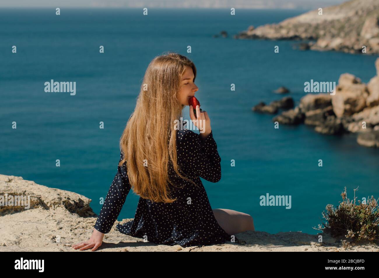 Belle fille blonde avec des yeux fermés mangeant la fraise et assis sur le bord de la montagne avec vue sur la mer, vue latérale Banque D'Images